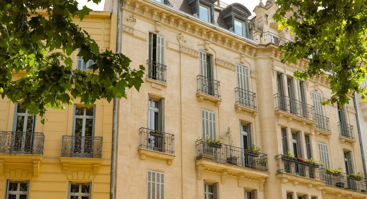 Elegant French-style architecture featuring balconies and detailed stonework under tree shade.