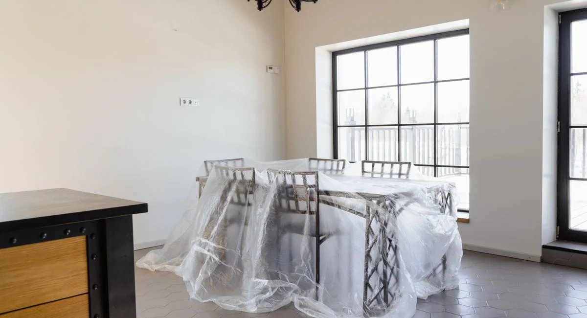 Bright dining room with table and chairs wrapped in protective plastic for renovation.
