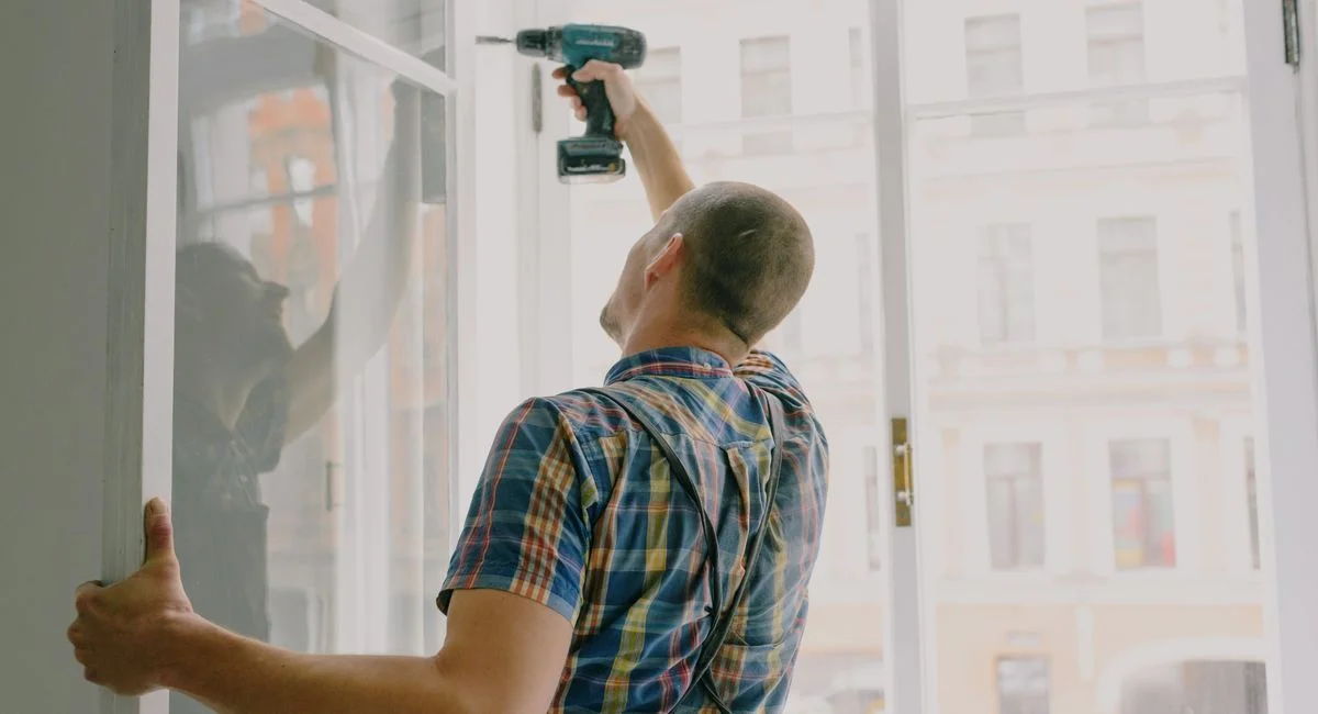 Back view of worker in checkered shirt drilling window frame during renovation process in apartment