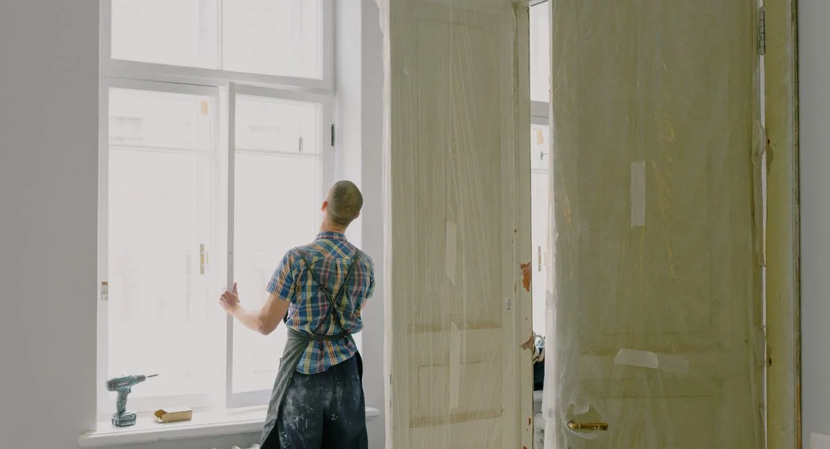 Back view of unrecognizable workman in apron mounting window near drilling machine and doors in flat