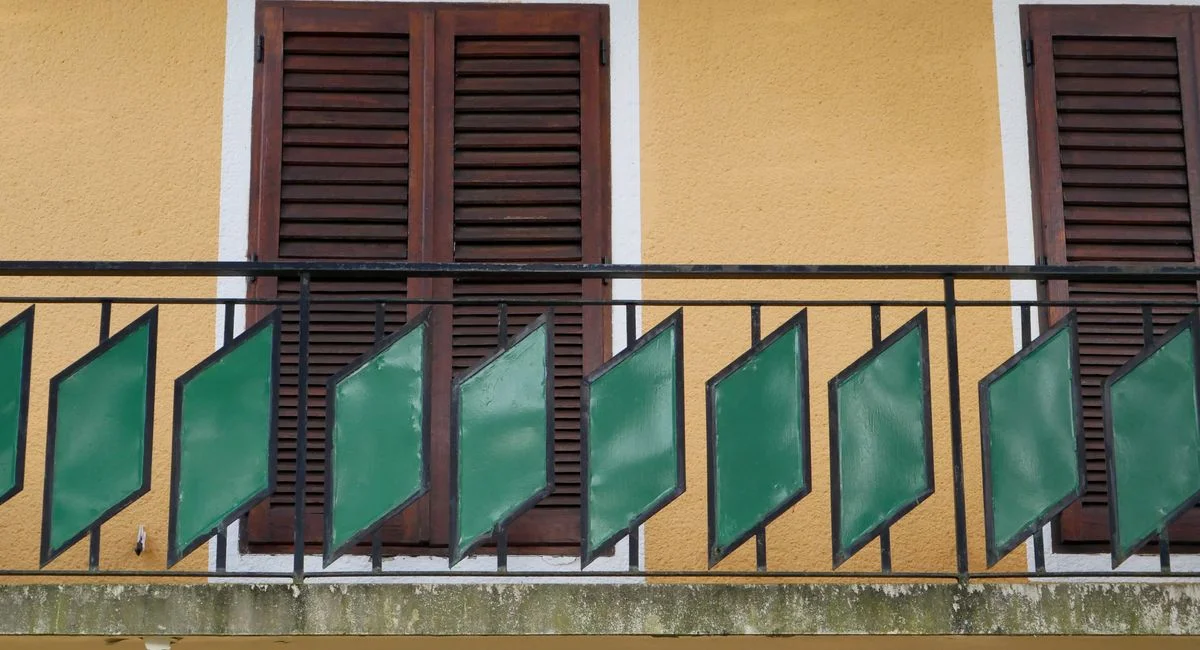 A vibrant yellow wall featuring a balcony with green geometric railing and shuttered windows.