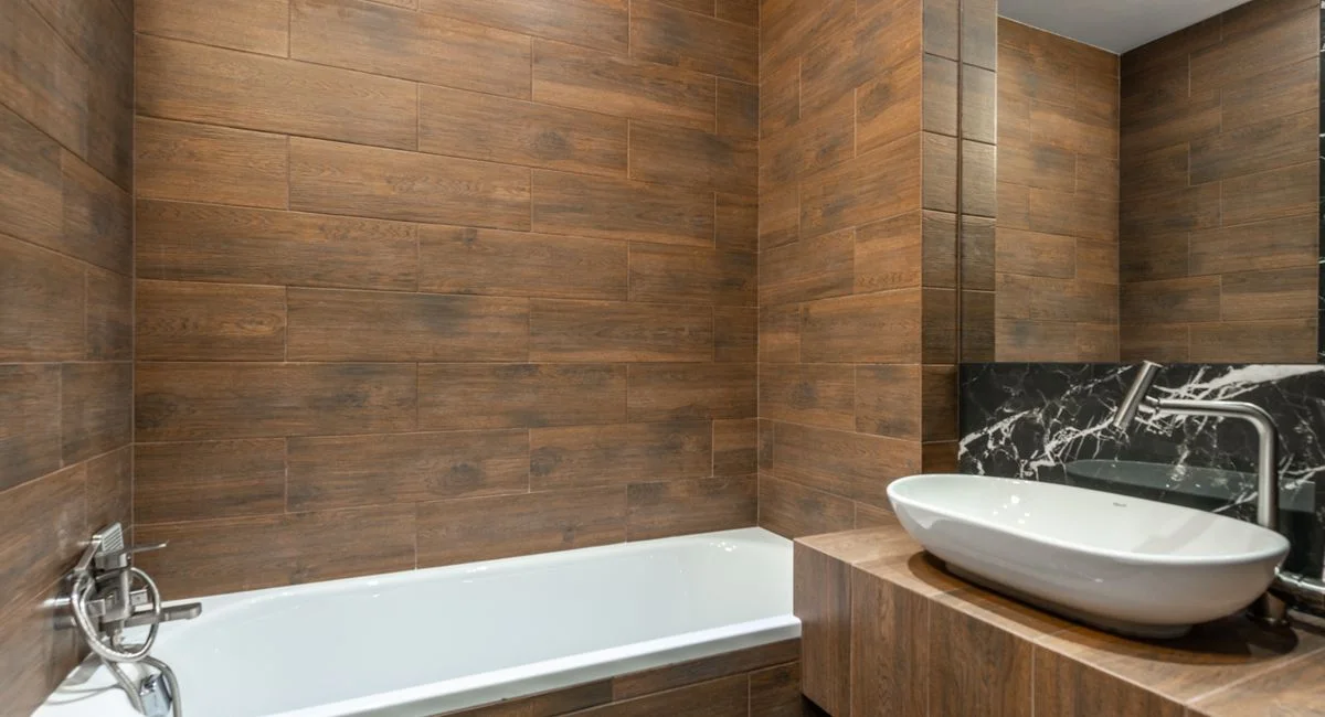 Stylish interior design of modern bathroom with wood like tiles equipped with white bathtub and sink with faucet