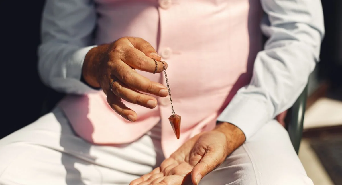 A man in soft pastel attire holding a gemstone pendulum over his palm outdoors.