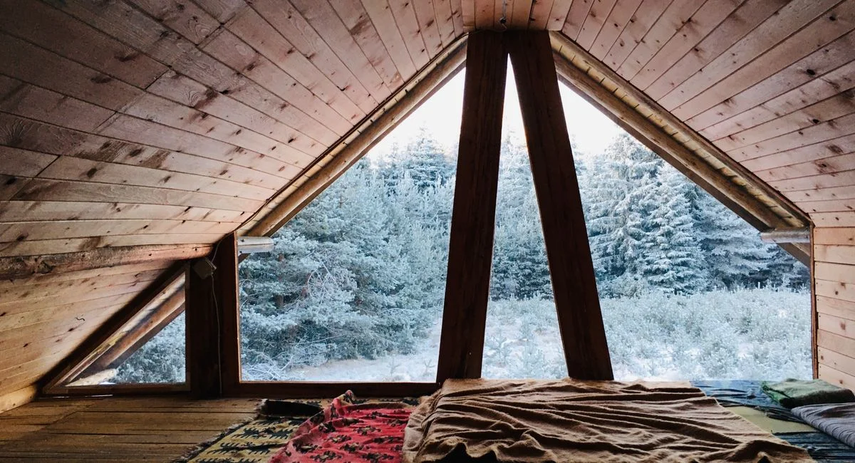 Warm attic room in wooden cabin with blankets overlooking a snowy forest.
