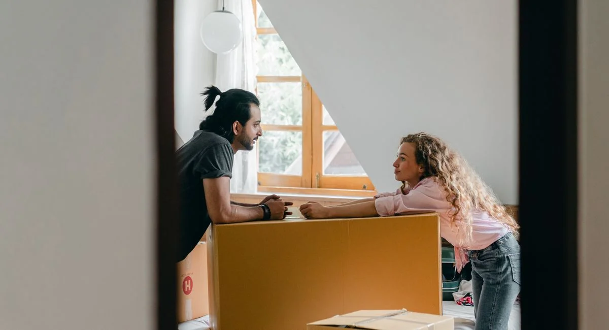 Side view from entrance of cheerful young ethnic bearded man with ponytail and woman with curly hair leaning on large cardboa