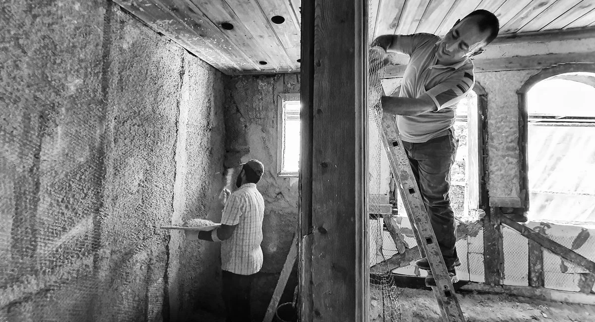 Two men renovating a room, one on a ladder applying plaster, showcasing teamwork and effort.