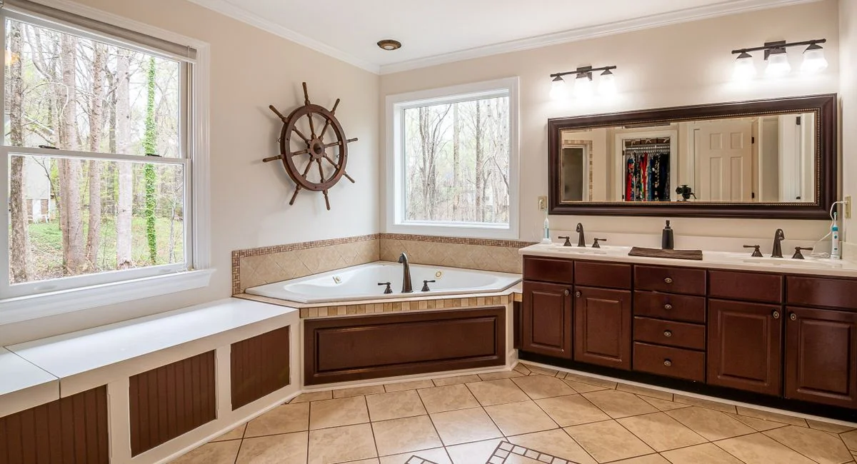 Spacious bathroom featuring a large corner bathtub, wooden cabinets, and natural light from large windows.