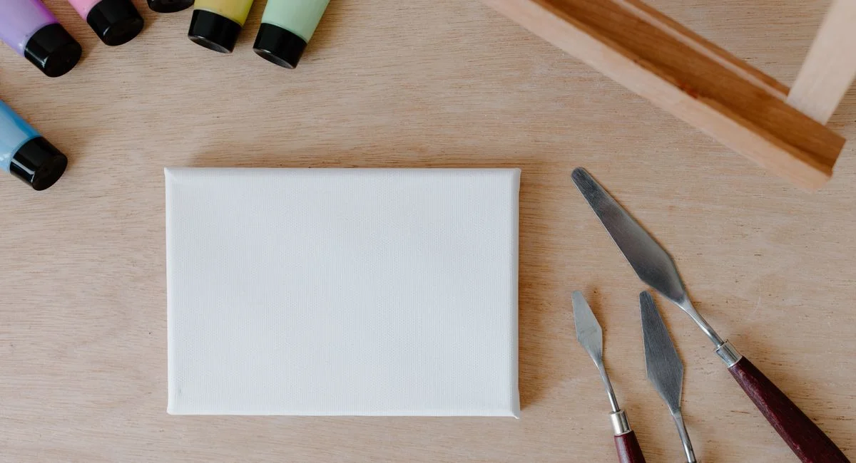 Top view of art supplies including paints, palette knives, and canvas on wooden surface.