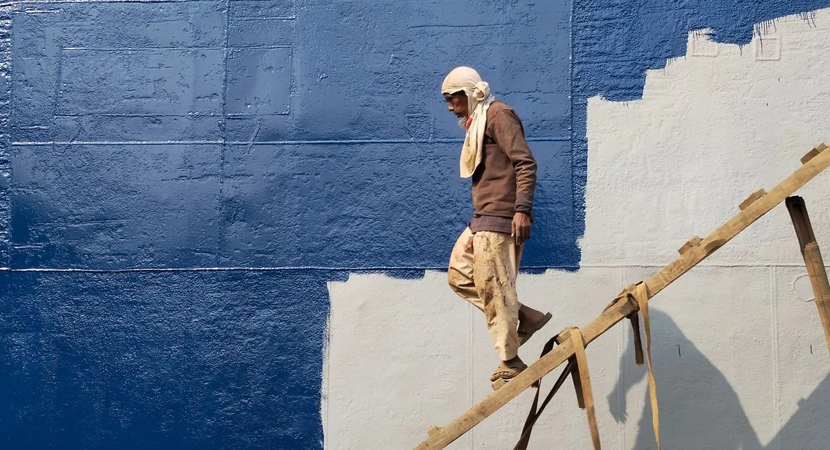 Candid street photo of a worker descending a ladder against a painted wall in Dhaka.
