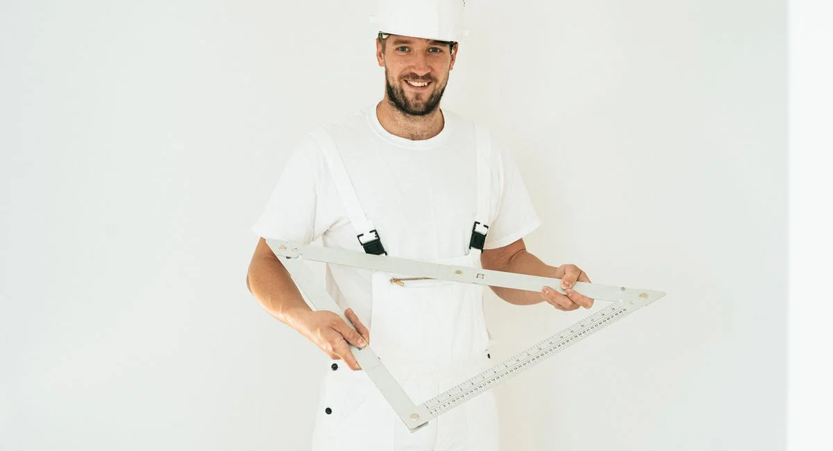 Happy carpenter in white outfit holding a large square ruler indoors.