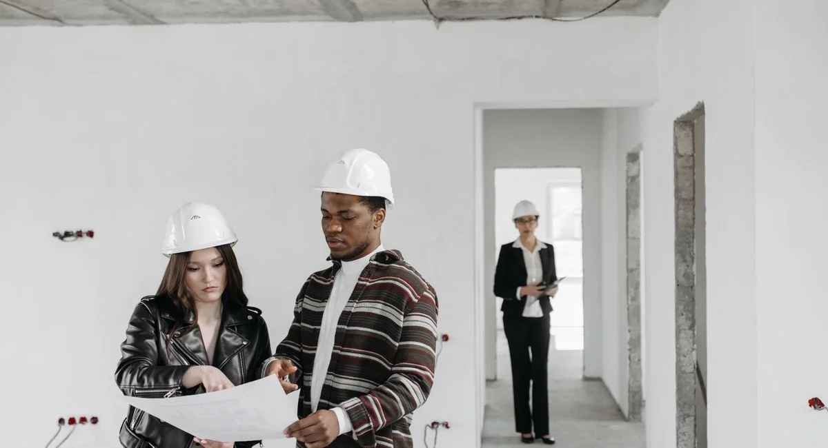 A couple wearing hard hats examines building plans inside an unfinished property with a realtor observing.